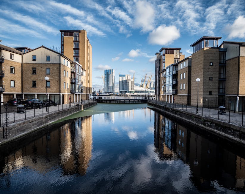 A scenic view of Docklands with a wooden pier in the foreground extending over calm water, featuring weathered wooden posts and planks. In the middle distance, there is a modern white building with te