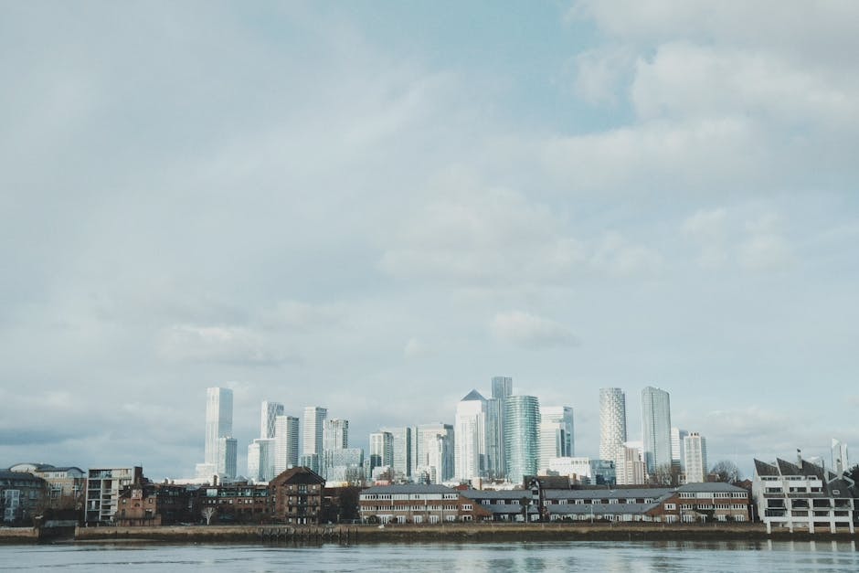A daytime view of a cityscape featuring numerous high-rise buildings with glass facades, situated behind a row of low-rise brick and concrete residential buildings. The high-rises are part of a modern financial district, with some skyscrapers reflecting the cloudy sky above. In the foreground, a body of water is visible, with the urban landscape directly behind it. The scene captures the exterior environment where home relocation or furniture transport might be occurring, providing context for professional removals services such as those offered by Man and a Van Docklands. The overall composition emphasizes the contrast between the older, smaller residential structures and the contemporary office towers, illustrating a typical high-rise flats environment relevant to house removals in the Docklands area. Good natural lighting highlights both the buildings and the waterfront setting, making it suitable for describing detailed moving and packing processes for the Canary Wharf removals guide.