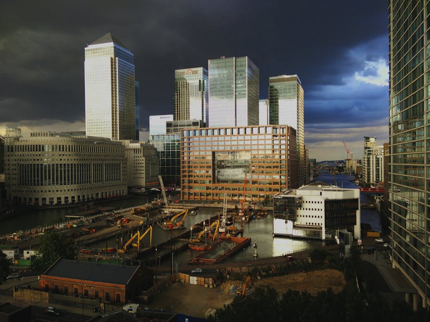 A daytime view of a modern urban waterfront area in Canary Wharf, Docklands, with several high rise office buildings made of glass and steel, some displaying reflections of the sky. In the foreground, there is a construction site with cranes, excavators, and construction vehicles, along with a temporary fence and construction materials. Nearby, a small brick building is visible on a street with parked cars. The scene is partially shaded by dark clouds, with patches of sunlight illuminating parts of the buildings and construction zone, capturing a moment of active building and development relevant to home relocation and furniture transport logistics assisted by Man and a Van Docklands.