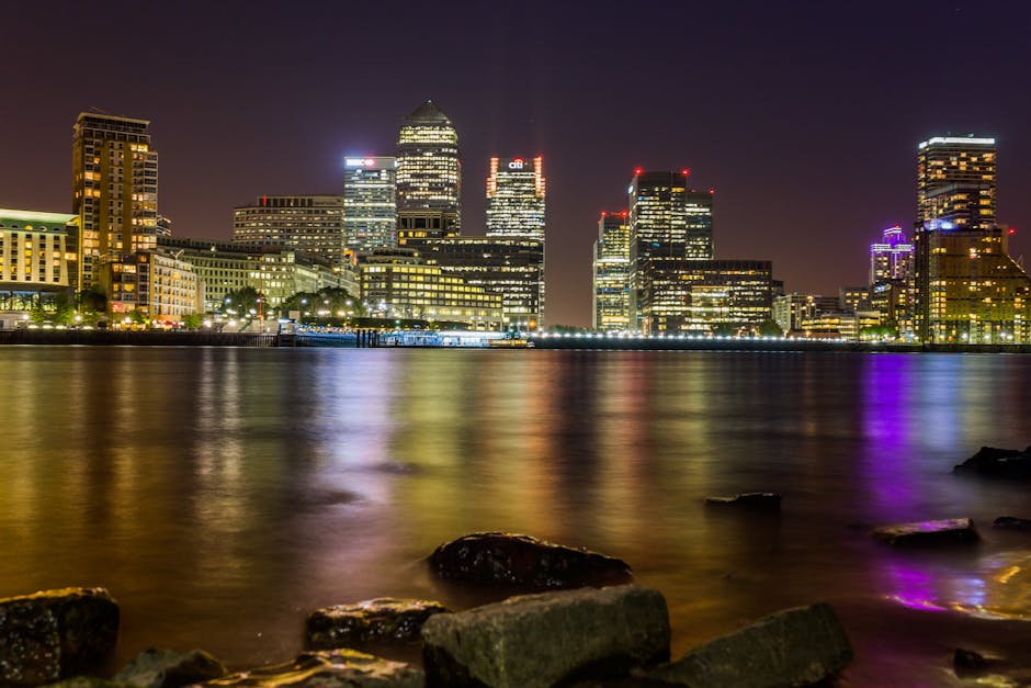 A daytime view of a cityscape featuring numerous high-rise buildings with glass facades, situated behind a row of low-rise brick and concrete residential buildings. The high-rises are part of a modern financial district, with some skyscrapers reflecting the cloudy sky above. In the foreground, a body of water is visible, with the urban landscape directly behind it. The scene captures the exterior environment where home relocation or furniture transport might be occurring, providing context for professional removals services such as those offered by Man and a Van Docklands. The overall composition emphasizes the contrast between the older, smaller residential structures and the contemporary office towers, illustrating a typical high-rise flats environment relevant to house removals in the Docklands area. Good natural lighting highlights both the buildings and the waterfront setting, making it suitable for describing detailed moving and packing processes for the Canary Wharf removals guide.