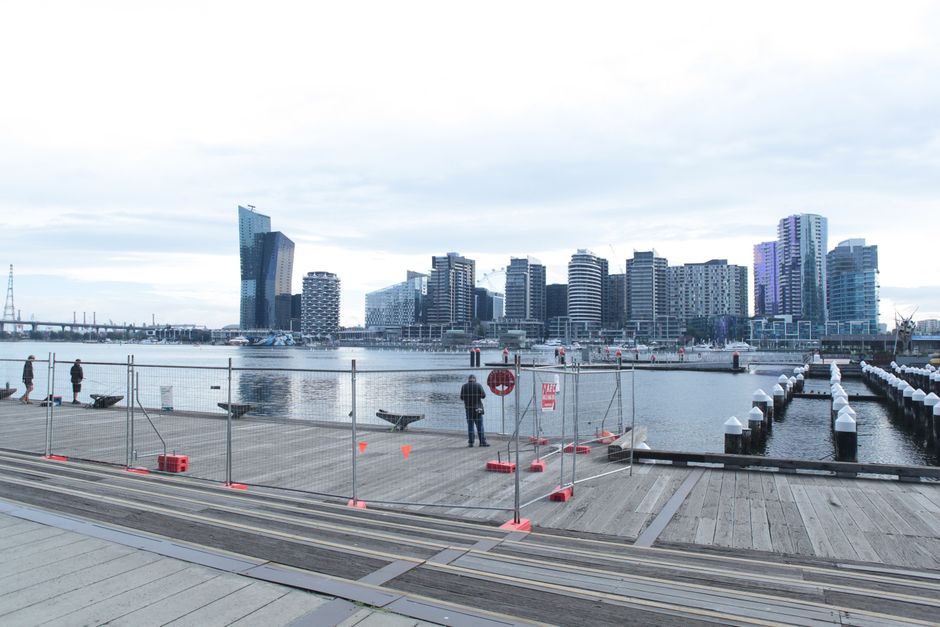 View of a waterfront dock area in Docklands on a cloudy day, featuring a wooden loading platform surrounded by temporary metal fencing with orange safety cones and signs. Several people are standing a