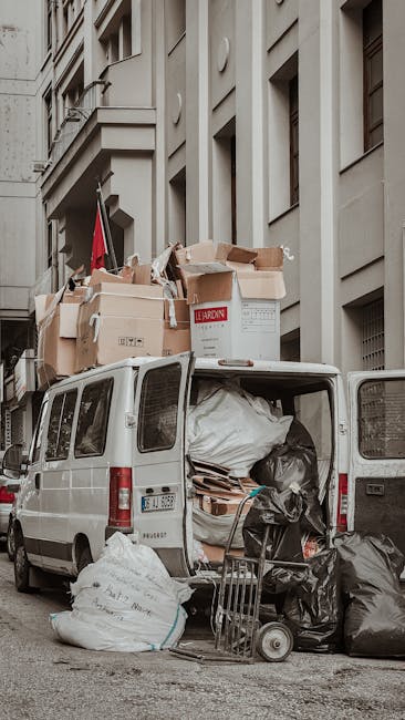 A white van parked on a narrow street in the Docklands area with its rear doors open, revealing a collection of packed cardboard boxes, some with visible labels and tape, along with large plastic bags filled with household items, all arranged inside and around the van for a home relocation. Several boxes are stacked on top of the van, secured with packing materials like bubble wrap and plastic wrapping. A small hand truck or dolly is positioned near the open van doors, ready for loading or unloading. The backdrop includes a tall, multi-storey building with concrete and stone facades, characteristic of urban residential or commercial architecture. The scene depicts the process of furniture transport and packing during a move, with natural daylight illuminating the objects and vehicle, consistent with the logistics of house removals in a dense city environment, as managed by companies like Man and a Van Docklands.