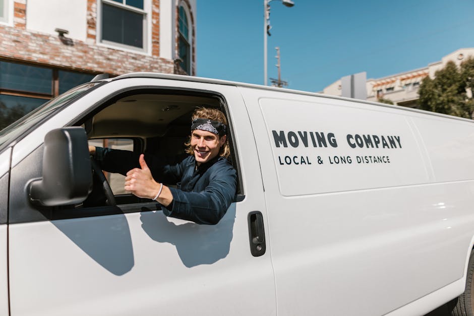 A young male mover with shoulder-length hair and wearing a headband is sitting in the driver’s seat of a white van labeled 'MOVING COMPANY LOCAL & LONG DISTANCE' on the side, inside a residential area suitable for home relocation. He is smiling and giving a thumbs-up through the open window, indicating readiness for a furniture transport or packing and moving process. The van is parked on a narrow street with brick and modern buildings visible in the background, under a clear blue sky. The scene includes part of the van's interior, showing a steering wheel and dashboard, with the mover positioned near the driver's side window. The environment suggests a professional removal and house moving service by Man and a Van Docklands, experienced in navigating tight urban streets for effective relocations.